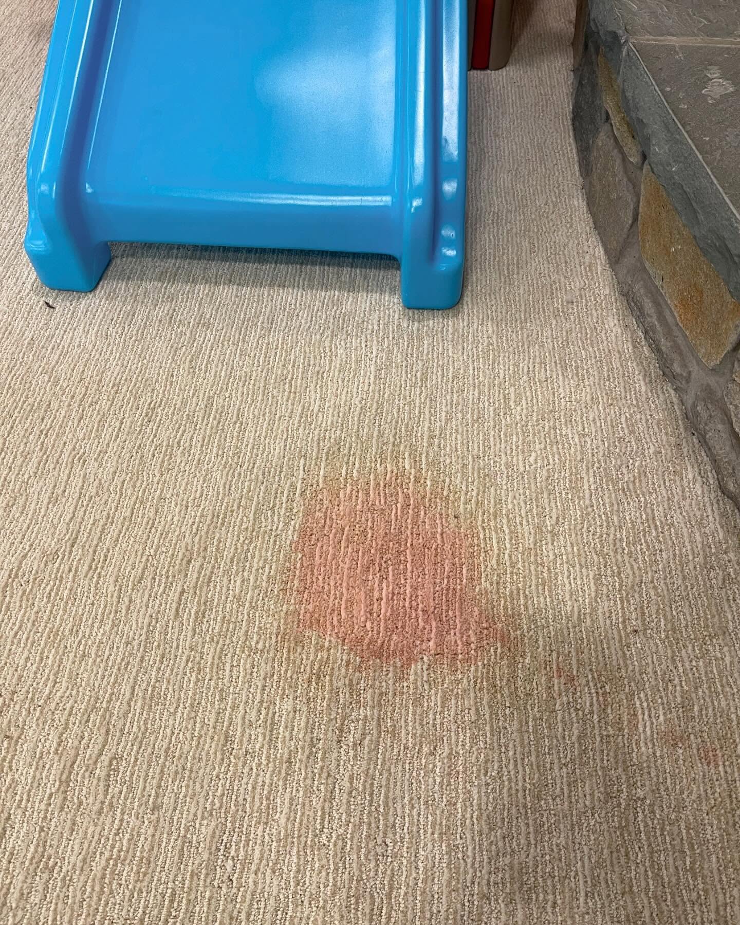 Large red juice dye stain on beige playroom carpet next to a blue slide